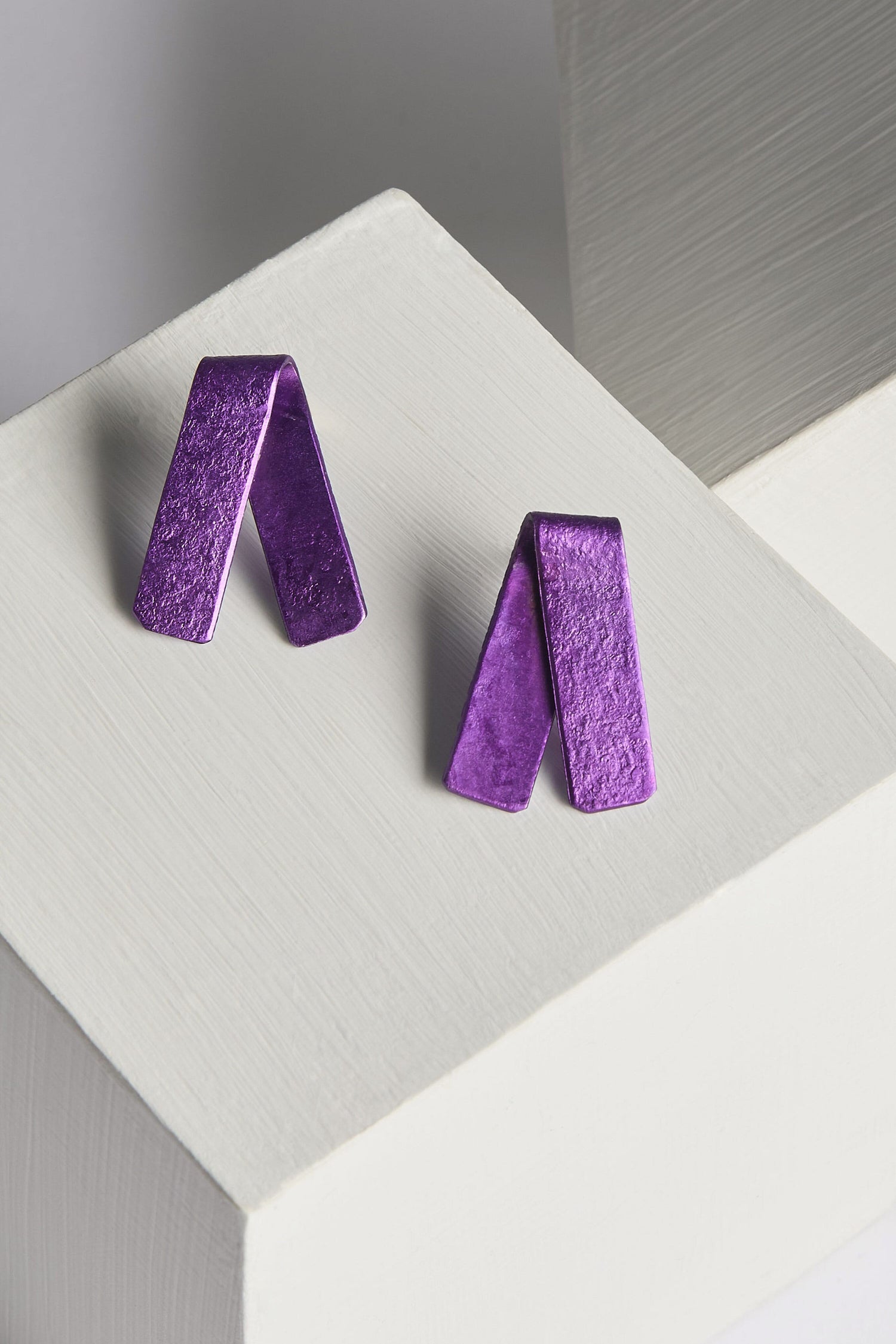 The Origami Earrings, a pair of vibrant, textured purple metal earrings shaped like folded ribbons and handmade by a Spanish design studio, are displayed on white geometric blocks.