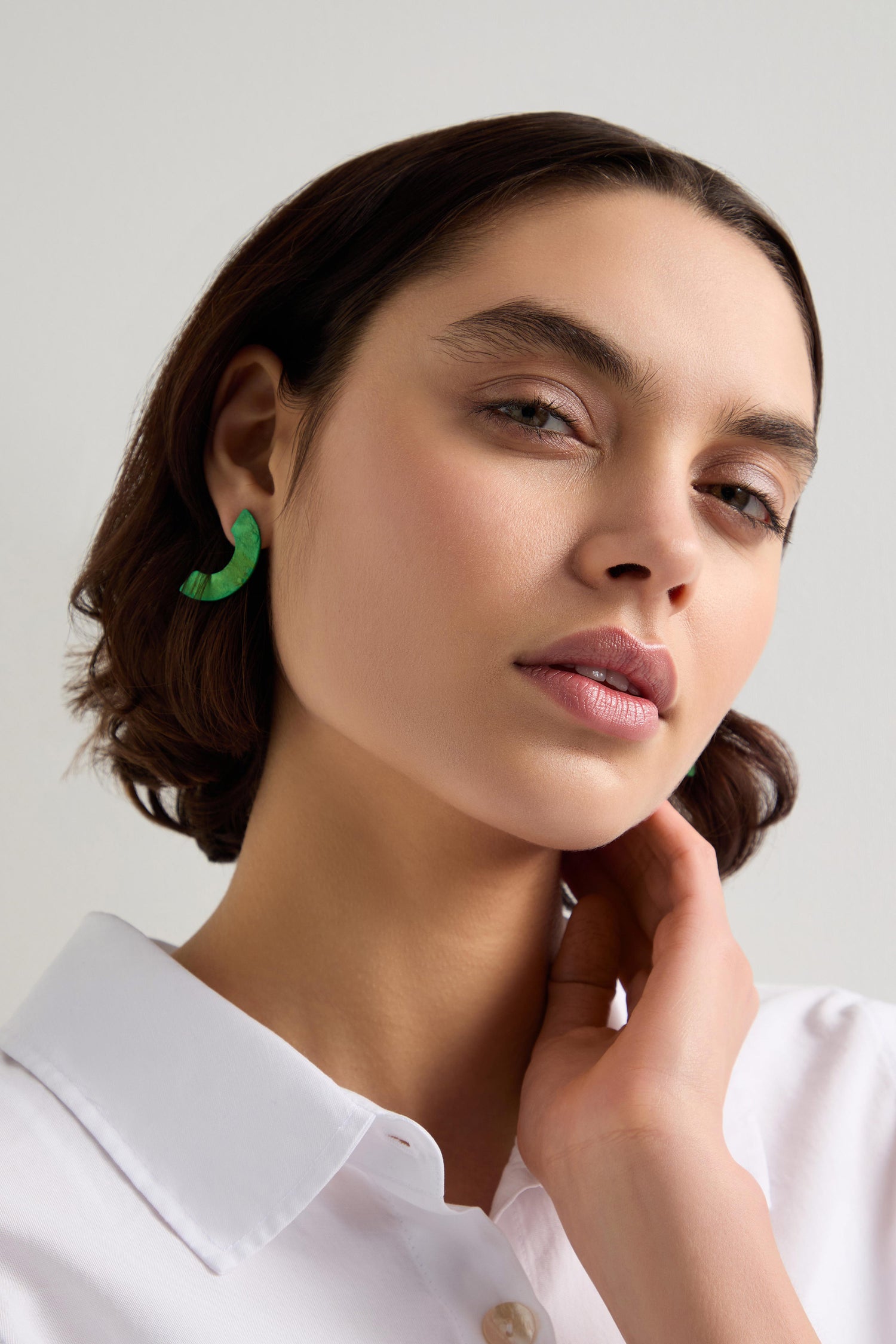 A person with short brown hair and standout accessories, featuring Aluminium Crescent Earrings, is wearing a white shirt. They are tilting their head slightly while touching their neck.