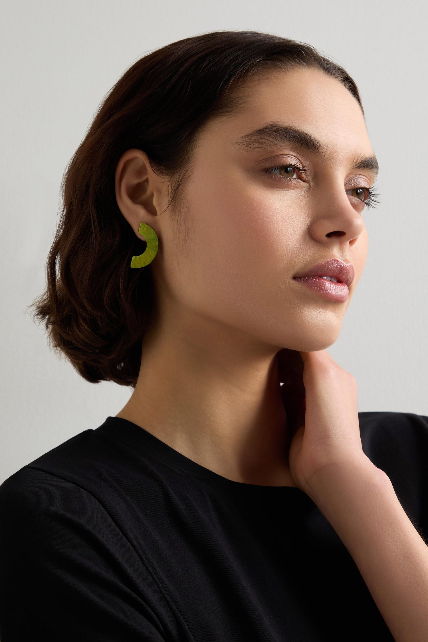 With short brown hair and green Aluminium Crescent Earrings, a person looks to the side, one hand near their neck, in a black top against a plain background.