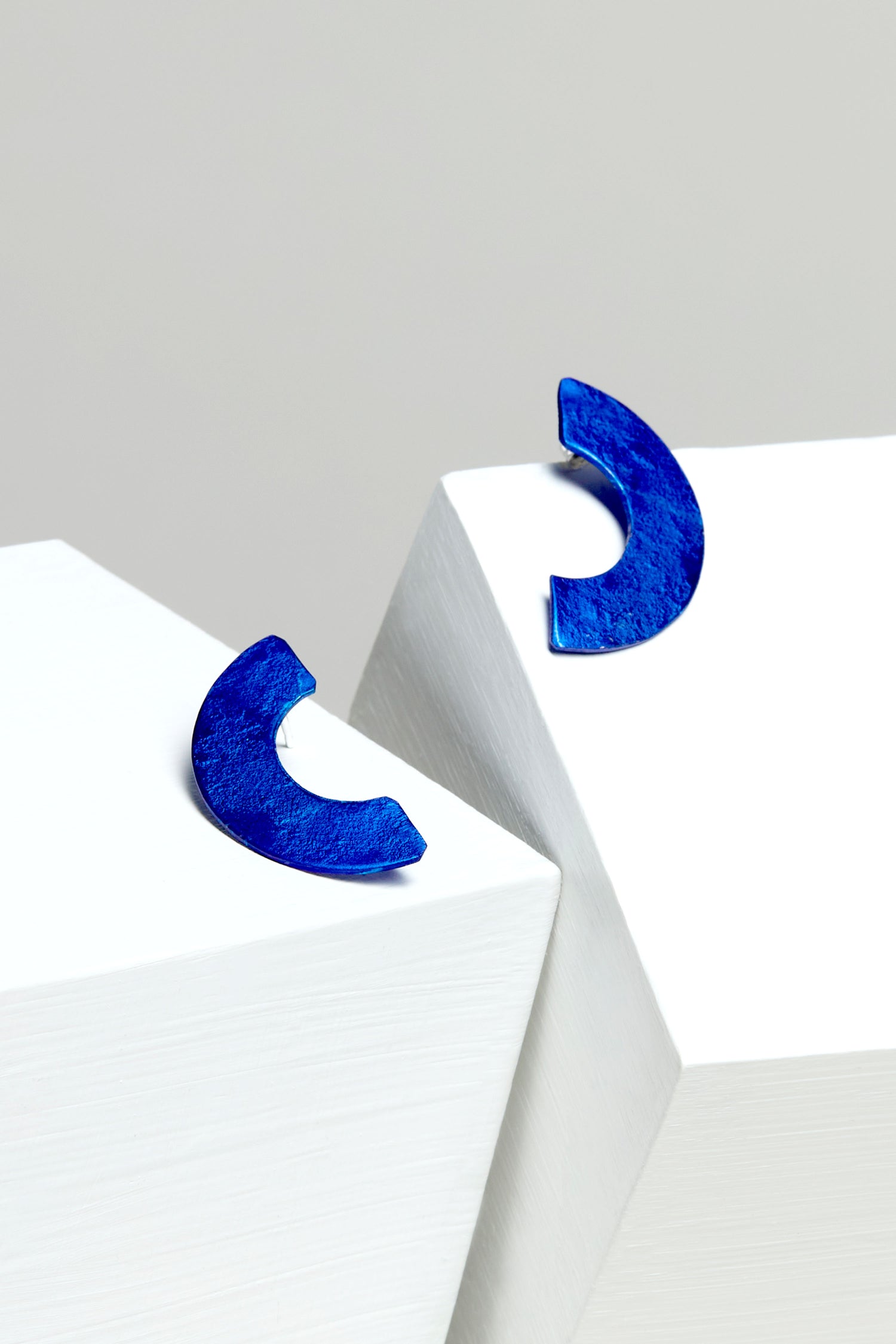 The elegant Aluminium Crescent Earrings, in blue, rest on angular white surfaces against a light gray background.