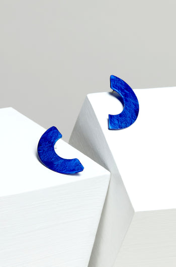 The elegant Aluminium Crescent Earrings, in blue, rest on angular white surfaces against a light gray background.