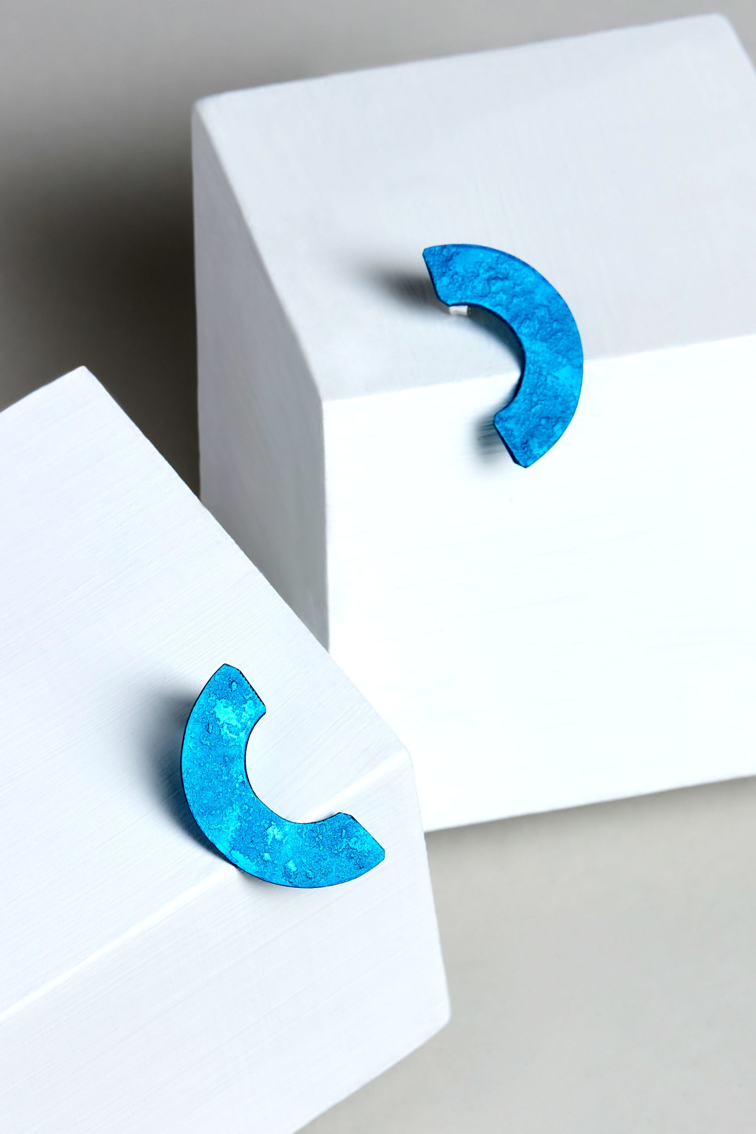 Two Aluminium Crescent Earrings, labeled OAE399, rest on white geometric blocks against a neutral background.