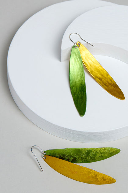 The Aluminium Leaves Earrings, in gleaming green and gold hues, rest elegantly on a white curved platform against a neutral backdrop. Perfect for adding nature-inspired glamour to any ensemble.