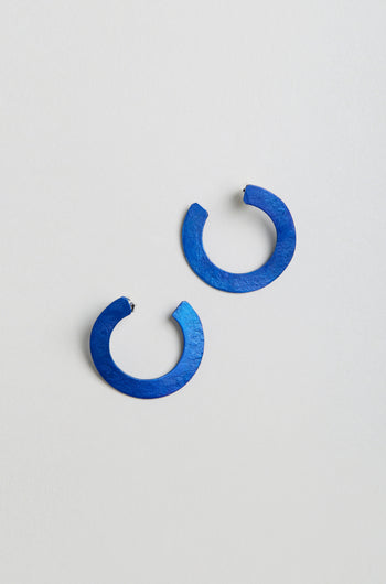 A pair of C Curve Earrings—round, flat, open-ended hoops in blue anodised aluminium—are displayed on a light grey surface.