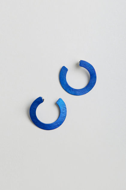 A pair of C Curve Earrings—round, flat, open-ended hoops in blue anodised aluminium—are displayed on a light grey surface.