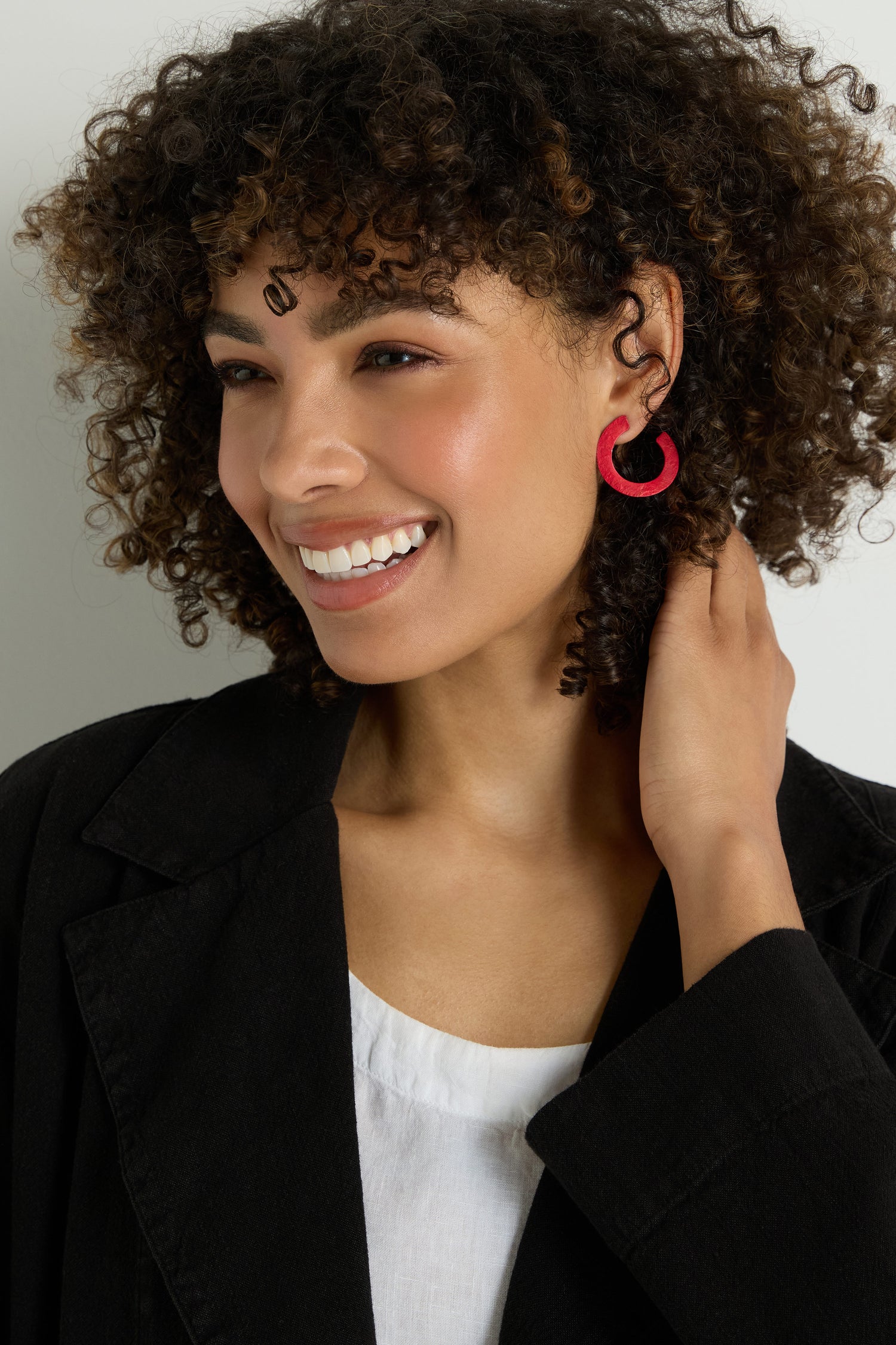 Smiling woman with curly hair wears a black jacket, white top, and C Curve Earrings, touching her hair with one hand.