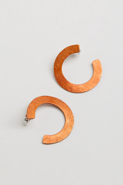 The C Curve Earrings feature a modern hoop shape in a distinctive C design, crafted from textured copper-colored metal and shown against a plain light background.