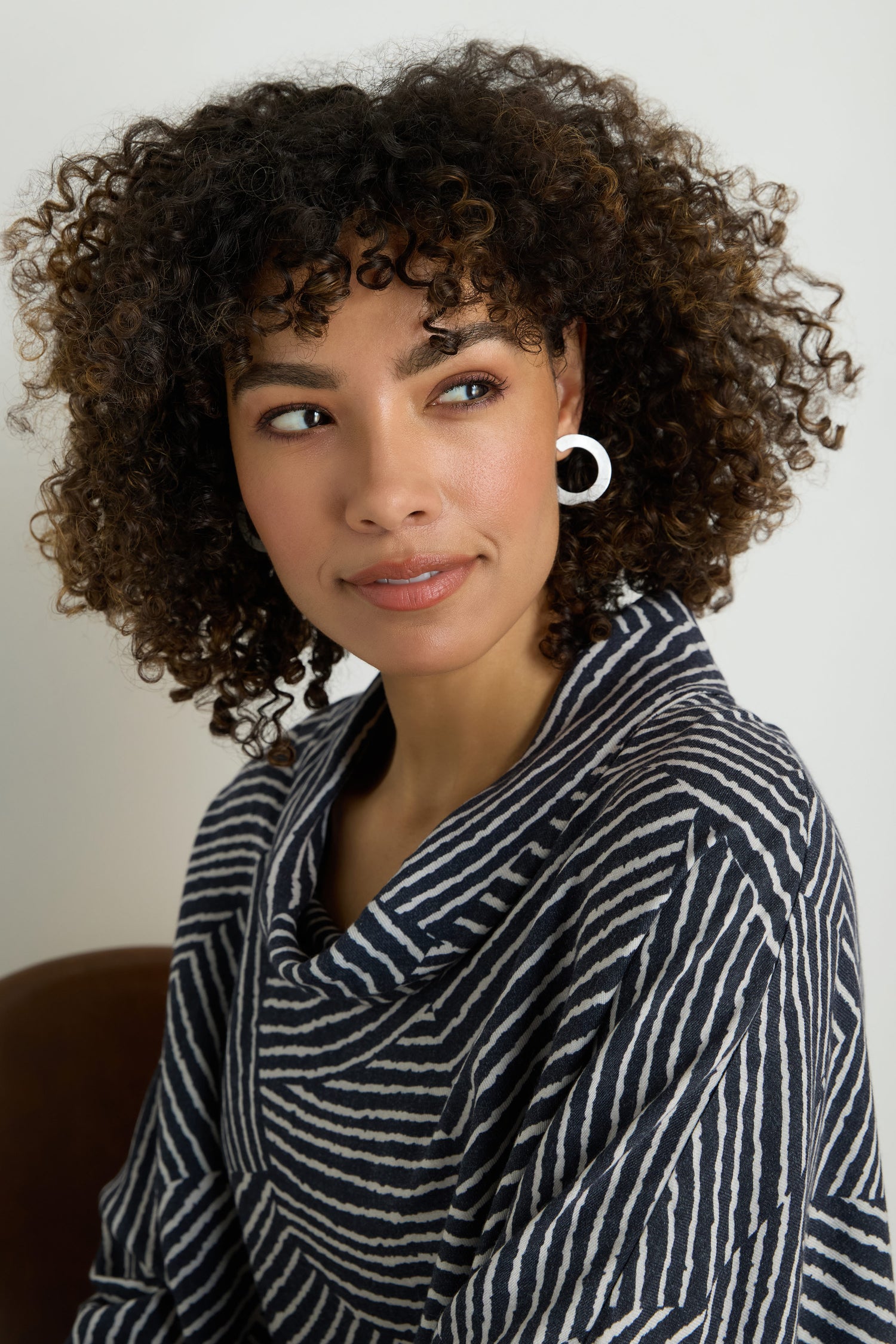 Woman with curly hair wears C Curve Earrings and a striped top, sitting and looking slightly to the side against a plain background.