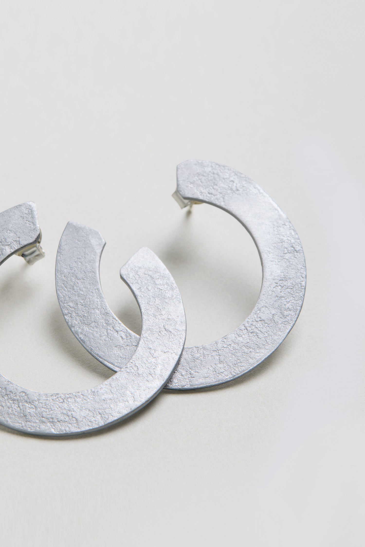 The C Curve Earrings feature a textured, silver-toned finish and a modern hoop design, displayed on a light-colored surface.