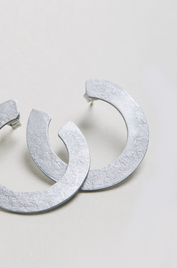 The C Curve Earrings feature a textured, silver-toned finish and a modern hoop design, displayed on a light-colored surface.