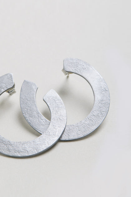 The C Curve Earrings feature a textured, silver-toned finish and a modern hoop design, displayed on a light-colored surface.