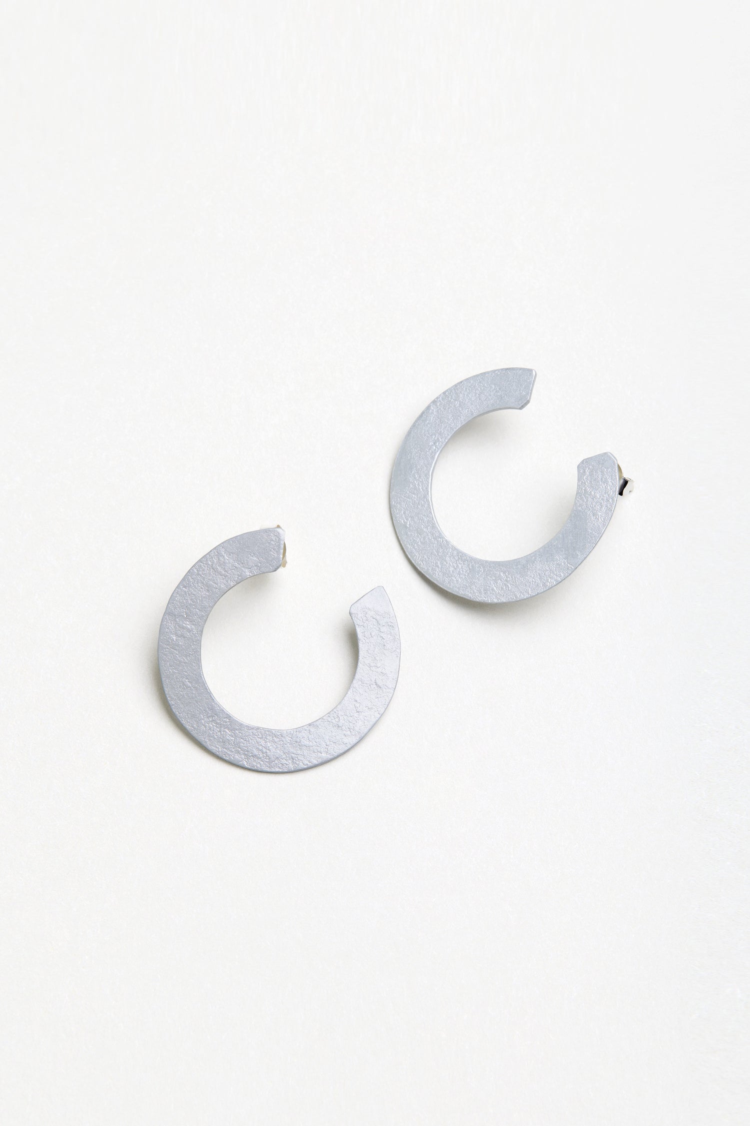 The C Curve Earrings are flat, silver-toned, modern hoops with an open circle design, shown against a plain white background.