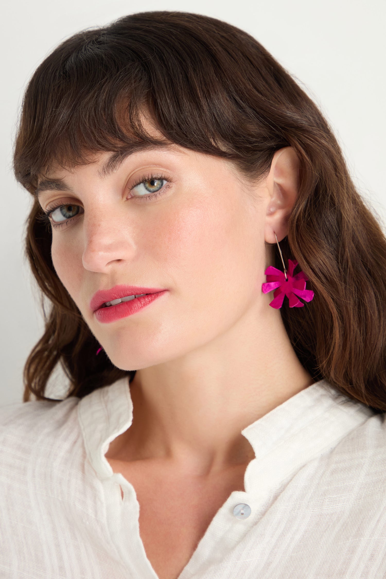 Woman with wavy brown hair wearing a white textured top and Flower Earrings—bright pink anodised aluminium flowers with sterling silver hooks—looking slightly to the side against a plain background.