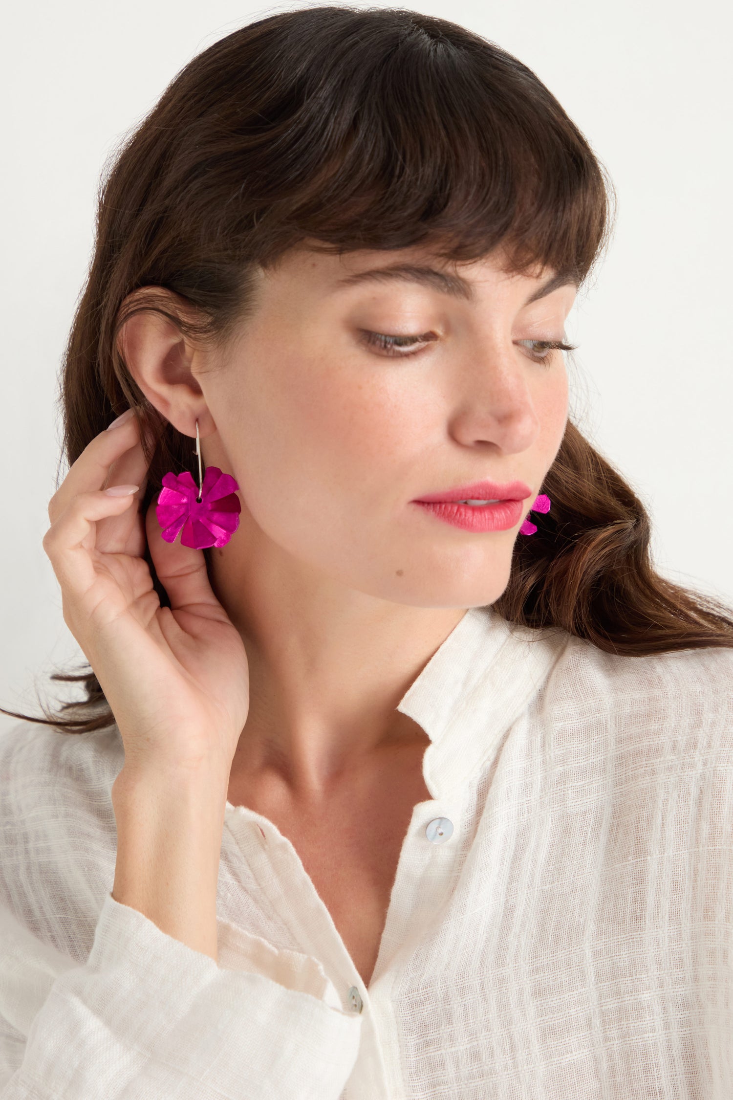 A woman looks down and touches one of her Flower Earrings—bright pink anodised aluminium flowers with sterling silver hooks—while wearing a white textured blouse.