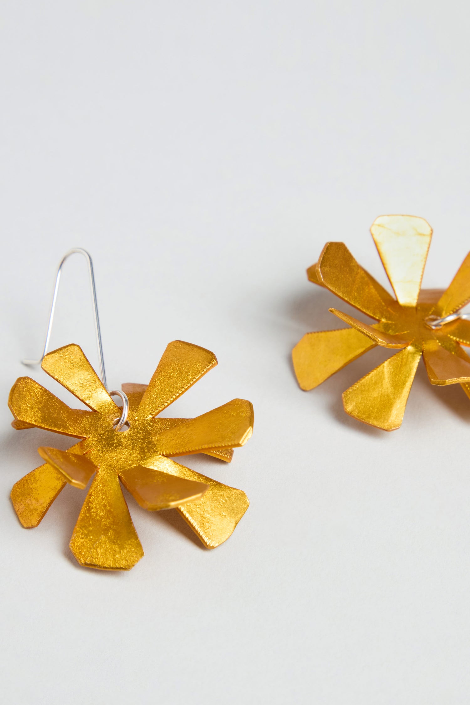 The Flower Earrings are gold-toned, flower-shaped metal earrings with petal-like segments and sterling silver hooks, displayed on a light background.