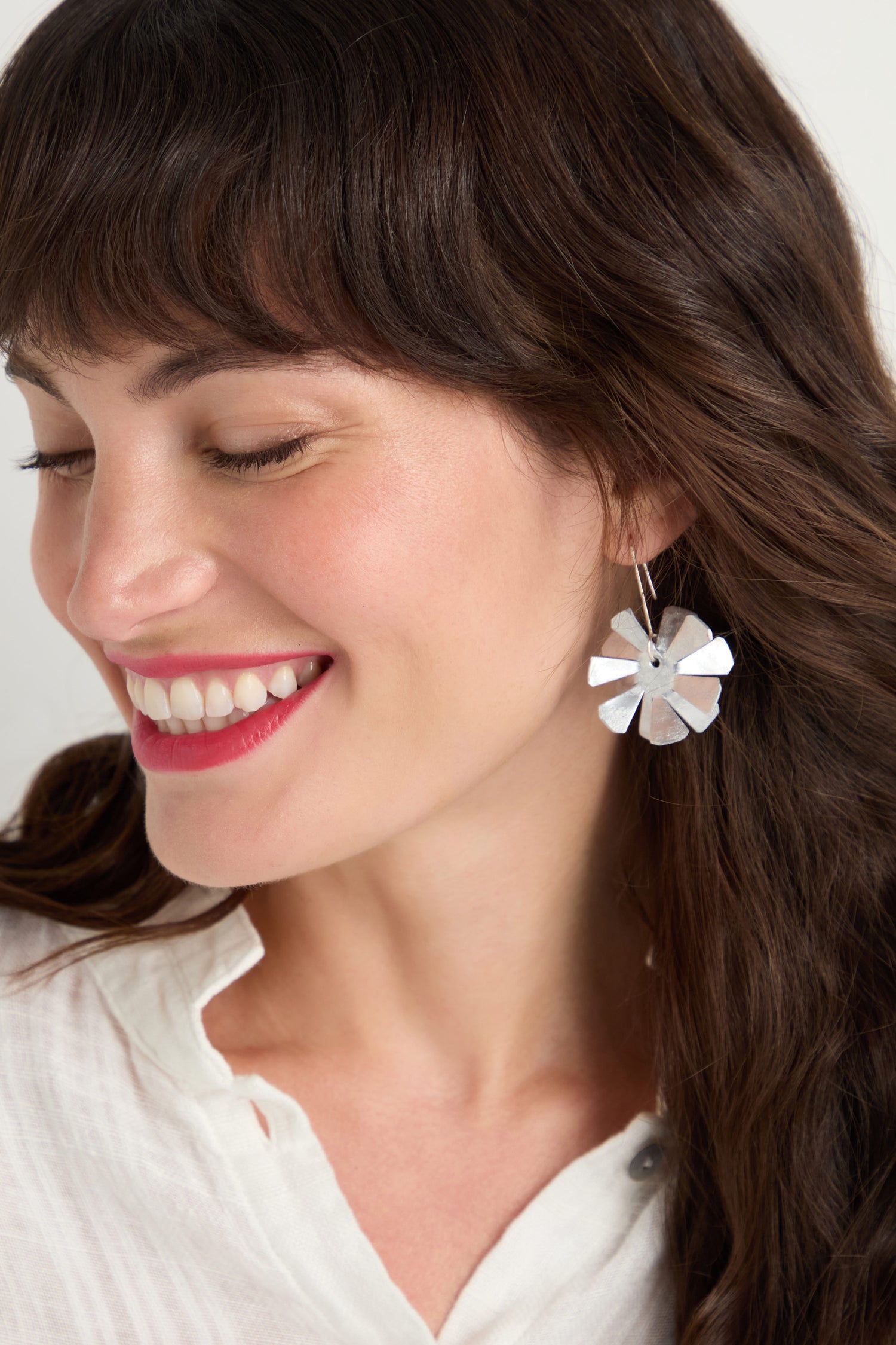 A woman with long brown hair smiles, wearing a white blouse and Flower Earrings made from anodised aluminium and finished with sterling silver hooks.