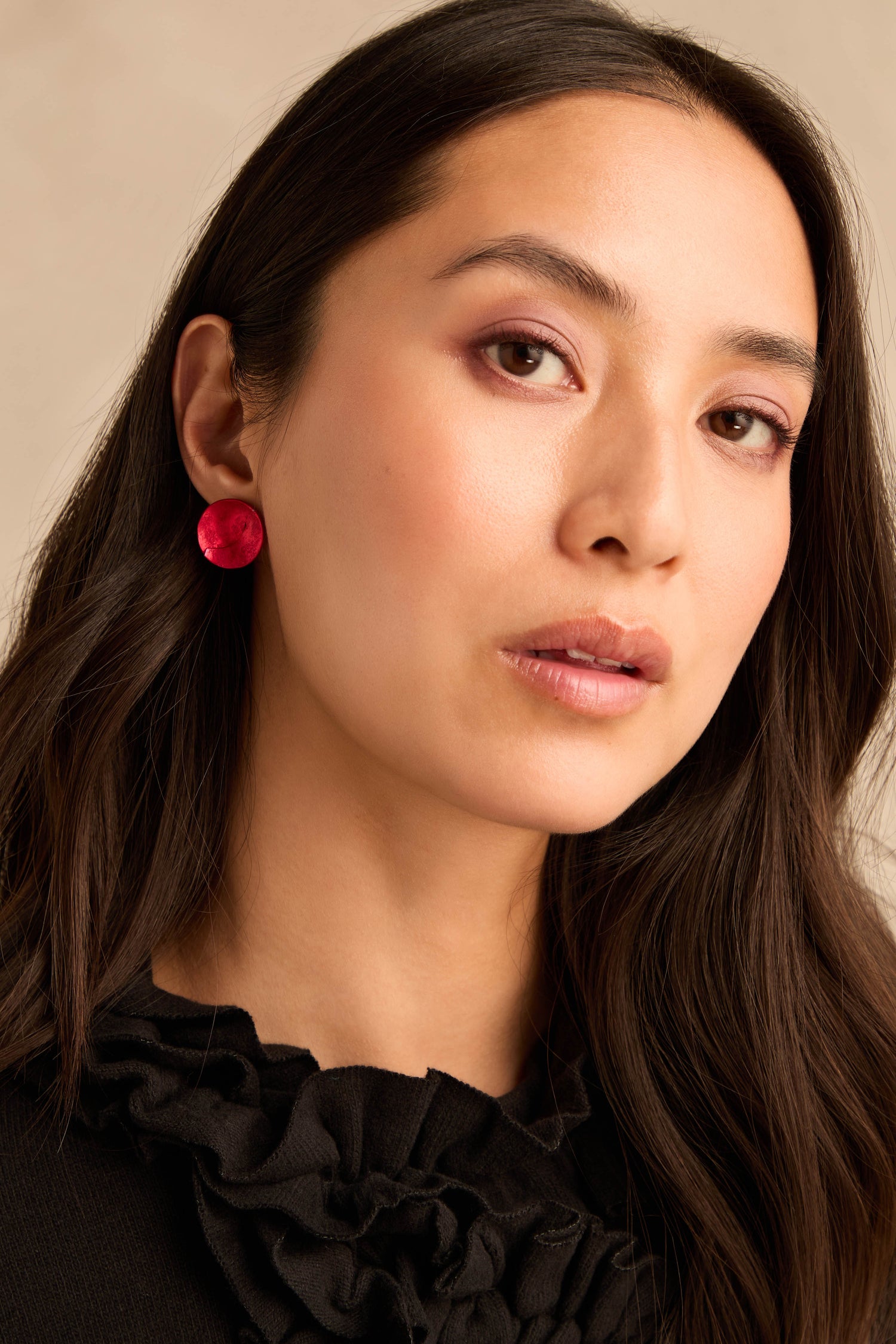A woman with long dark hair is wearing a black ruffled top and the Curved Disc Earrings—large pink anodised aluminium discs with sterling silver posts—while looking at the camera with a neutral expression.
