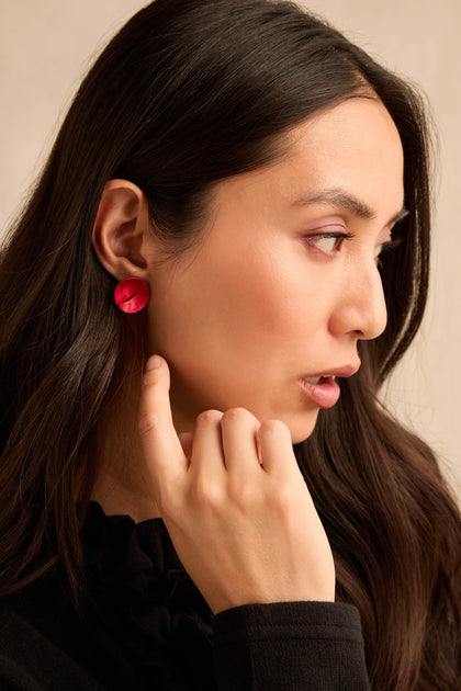 A woman with long dark hair wears striking red Curved Disc Earrings, crafted from anodised aluminium with sterling silver posts. She pairs them with a black top and looks to the right, her hand near her chin.