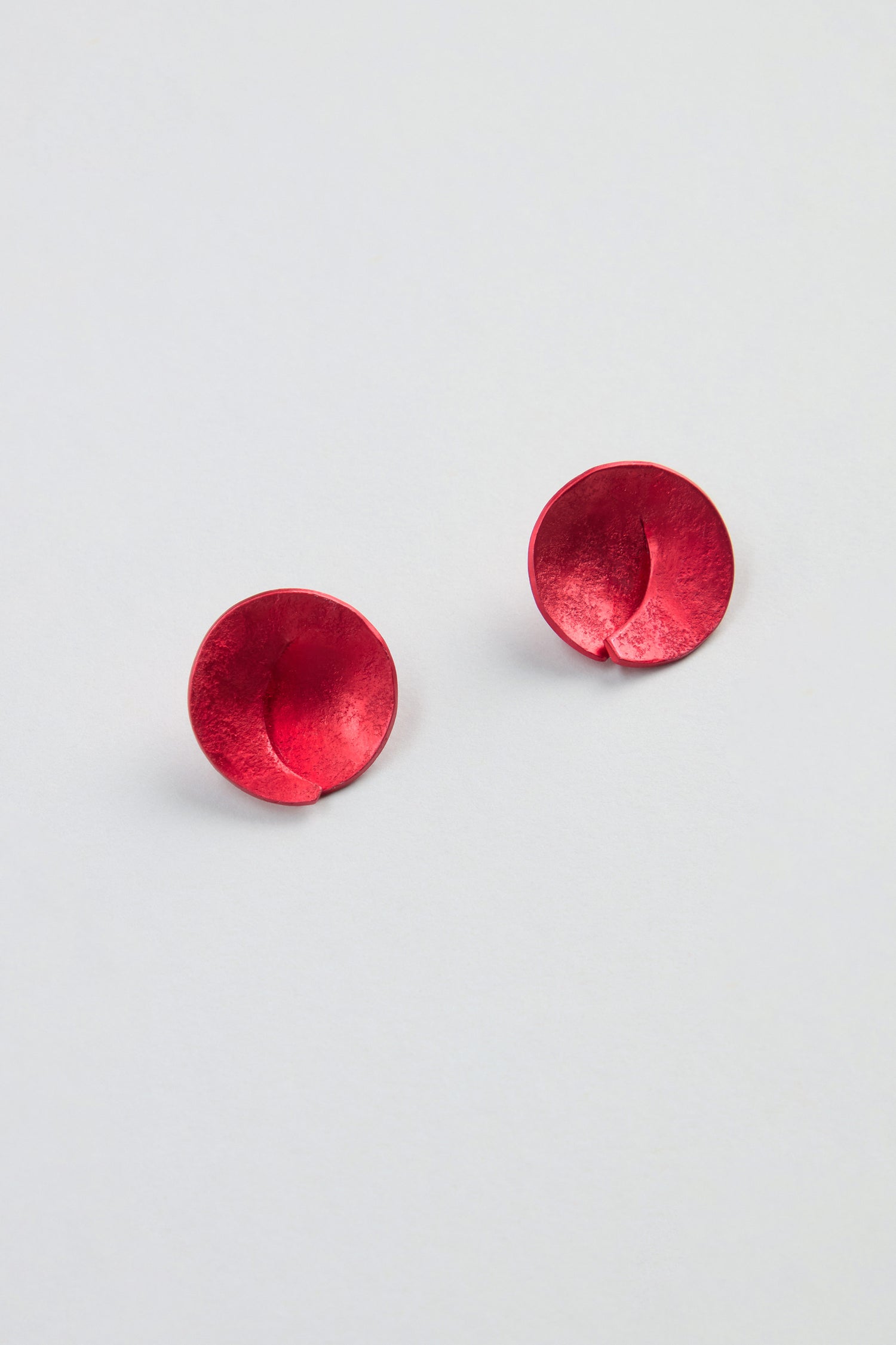 A pair of Curved Disc Earrings, featuring small, round, red discs with a slightly curved and overlapping design, finished with sterling silver posts and displayed on a simple light background.
