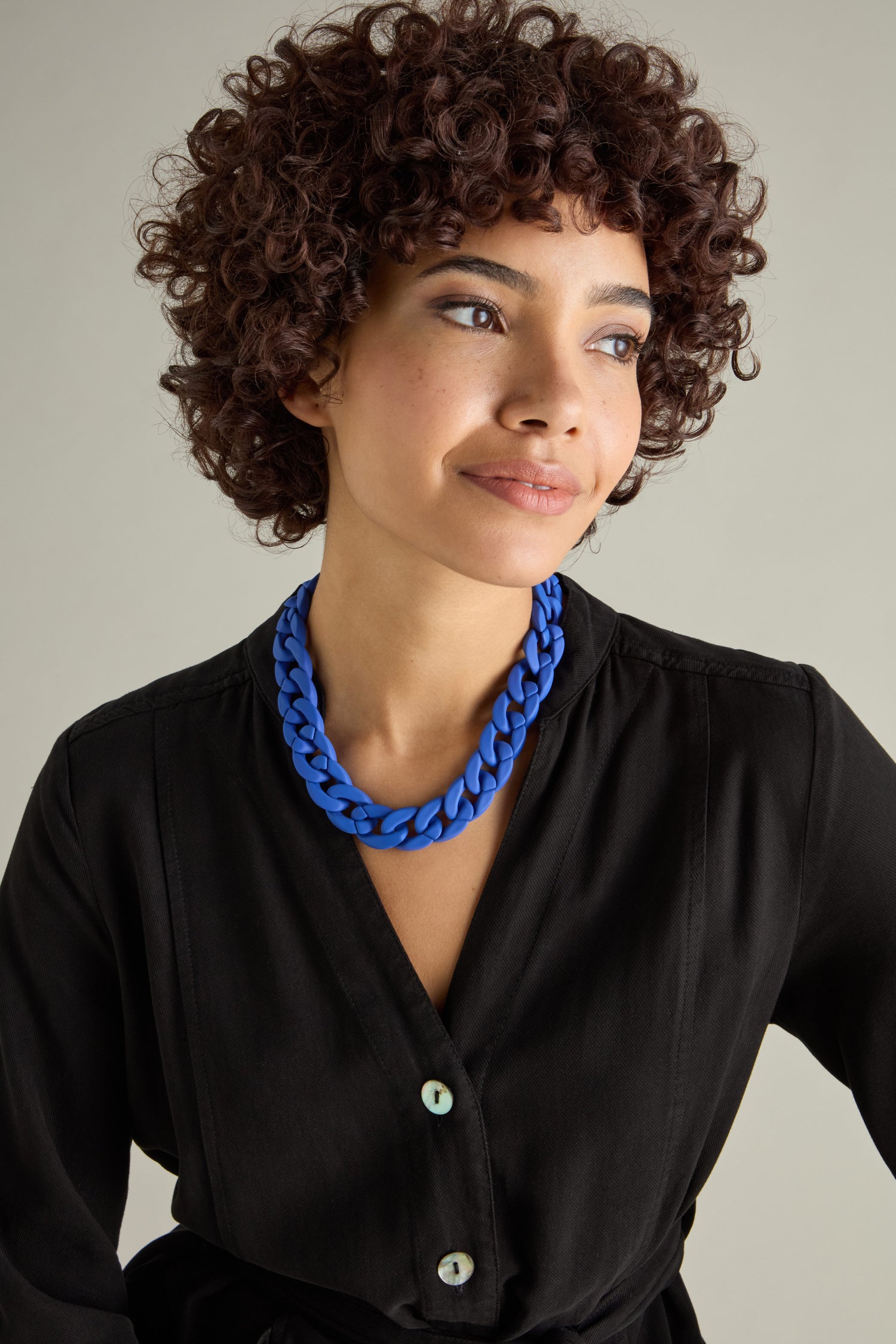 A curly-haired person wearing a black top and the bold Colour Pop Link Necklace looks to the side against a neutral background.