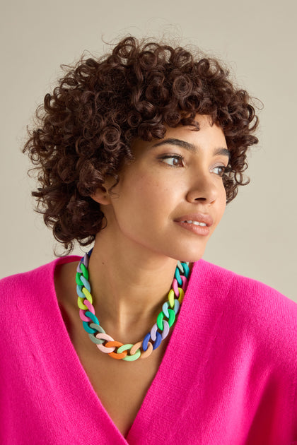 A person with curly hair wears a bright pink top and a bold Colour Pop Link Necklace, glancing to the side against a plain background.