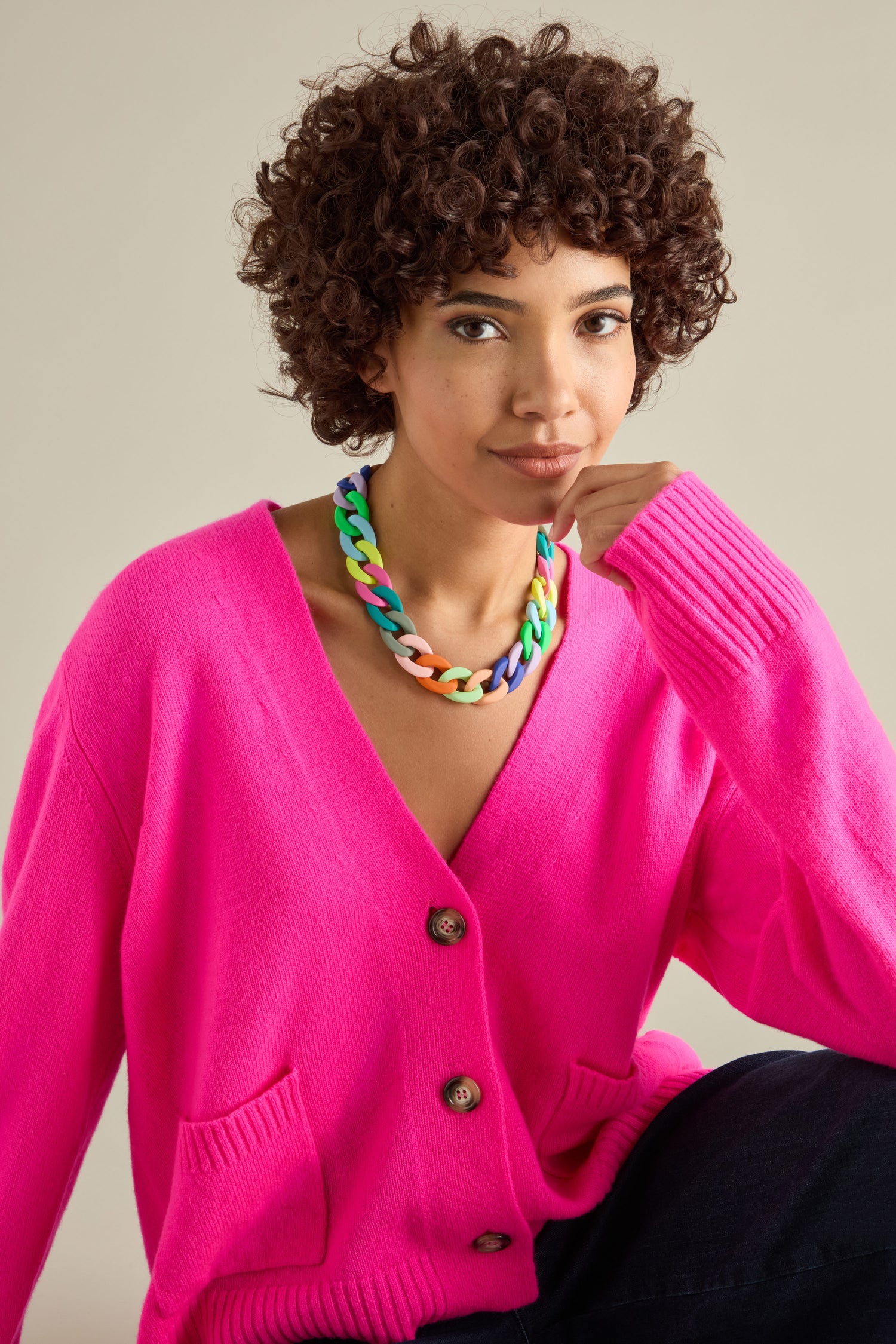 A person with curly hair is wearing a bright pink cardigan paired with a Colour Pop Link Necklace, showcasing bold accessories.