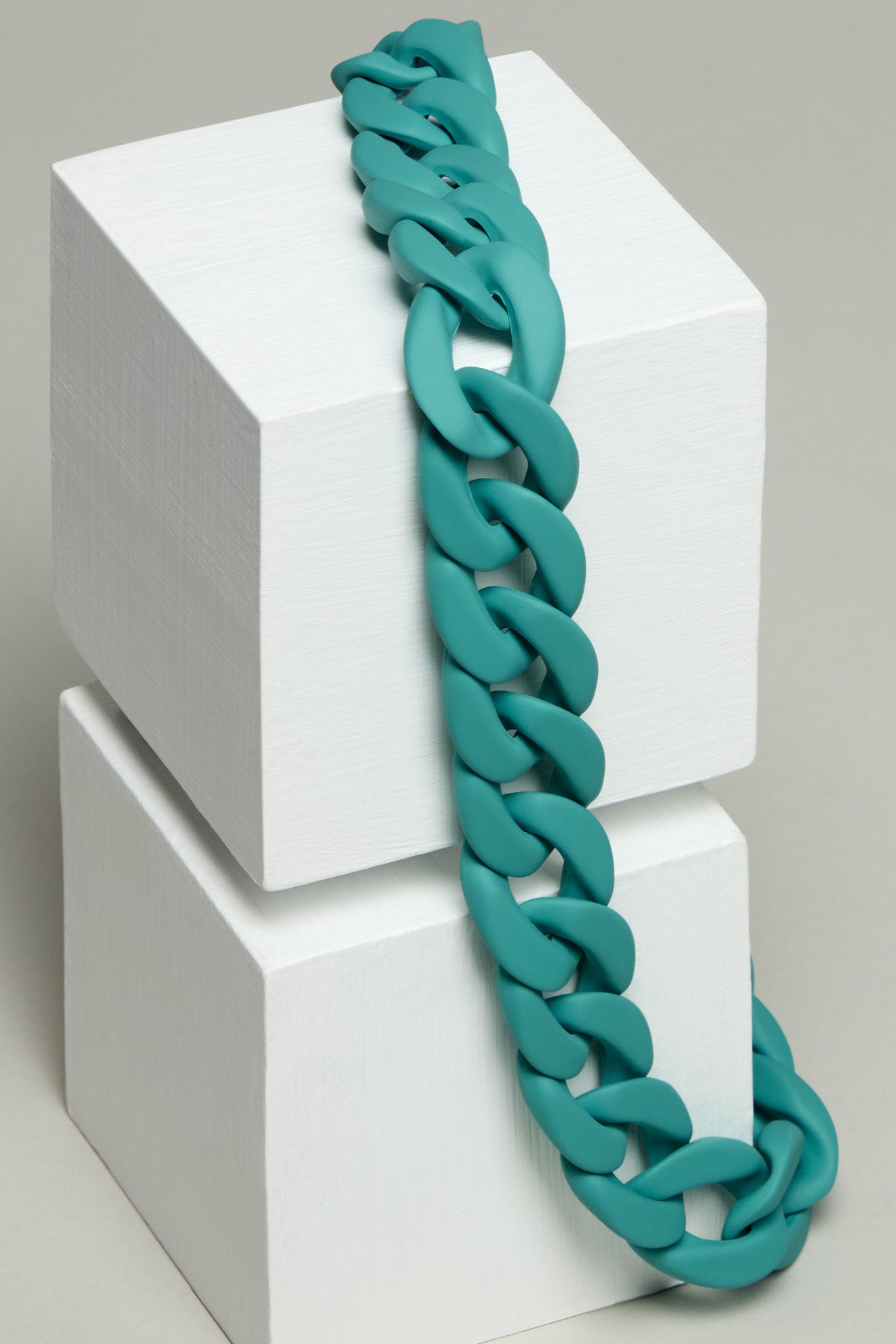 The Colour Pop Link Necklace, a teal chunky chain, draped over two stacked white cubes against a plain background.