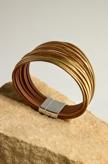 The Woven Metallic Bracelet, featuring a gold and brown multi-strand design with a metallic finish, is elegantly displayed on a beige textured rock, secured by a sleek silver clasp.