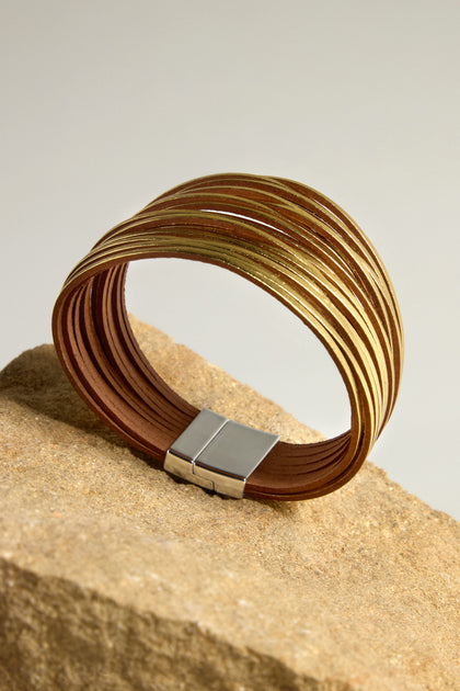 The Woven Metallic Bracelet, featuring a gold and brown multi-strand design with a metallic finish, is elegantly displayed on a beige textured rock, secured by a sleek silver clasp.