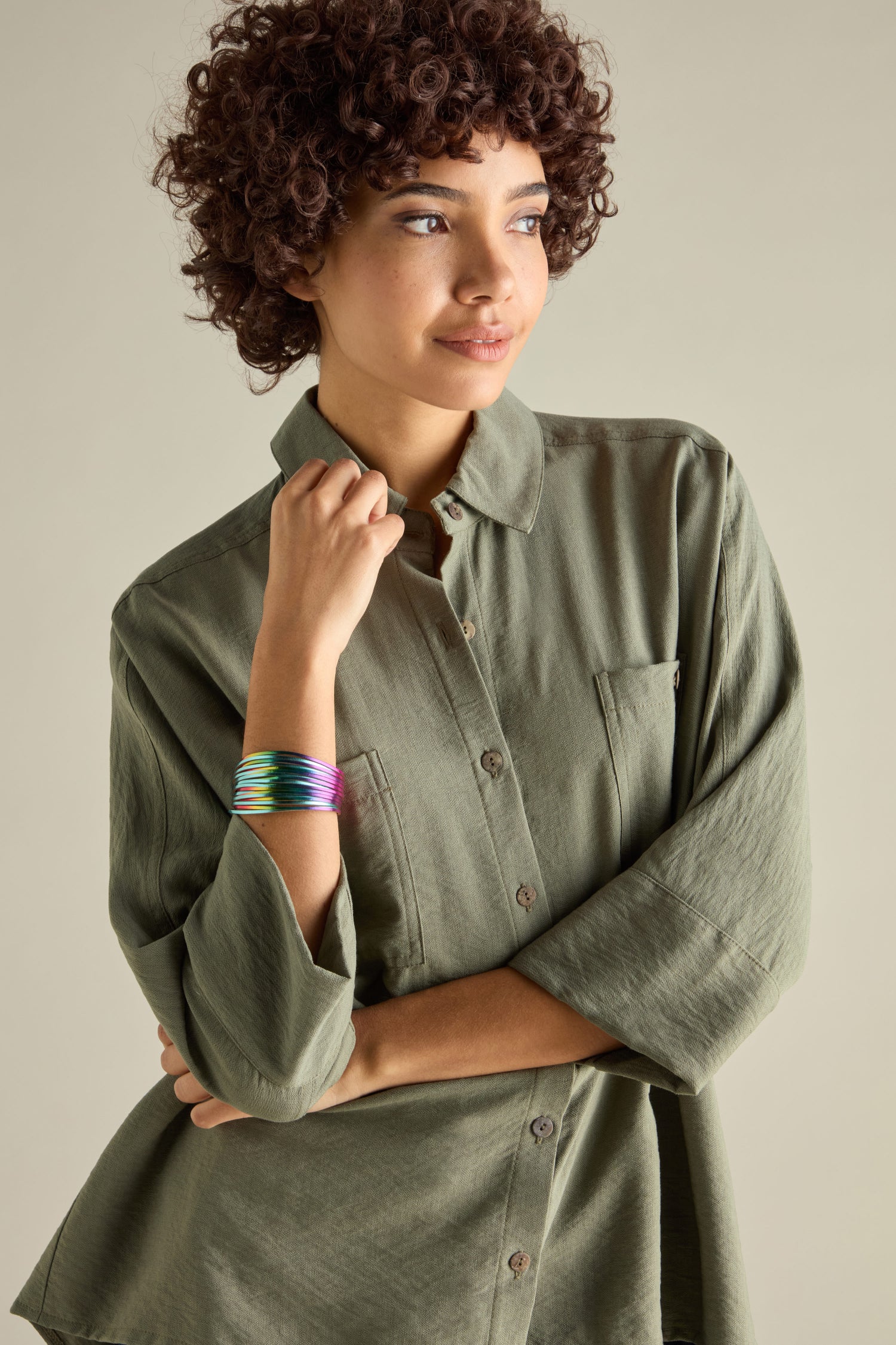 A person with curly hair in a green button-up shirt and a vibrant woven metallic bracelet stands with arms crossed, gazing to the side against a neutral background.