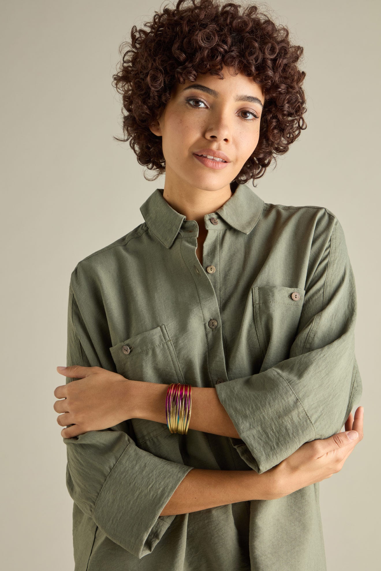 A curly-haired person in a green shirt stands with arms crossed, wearing a Woven Metallic Bracelet adorned with twisted leatherette and a magnetic clasp.