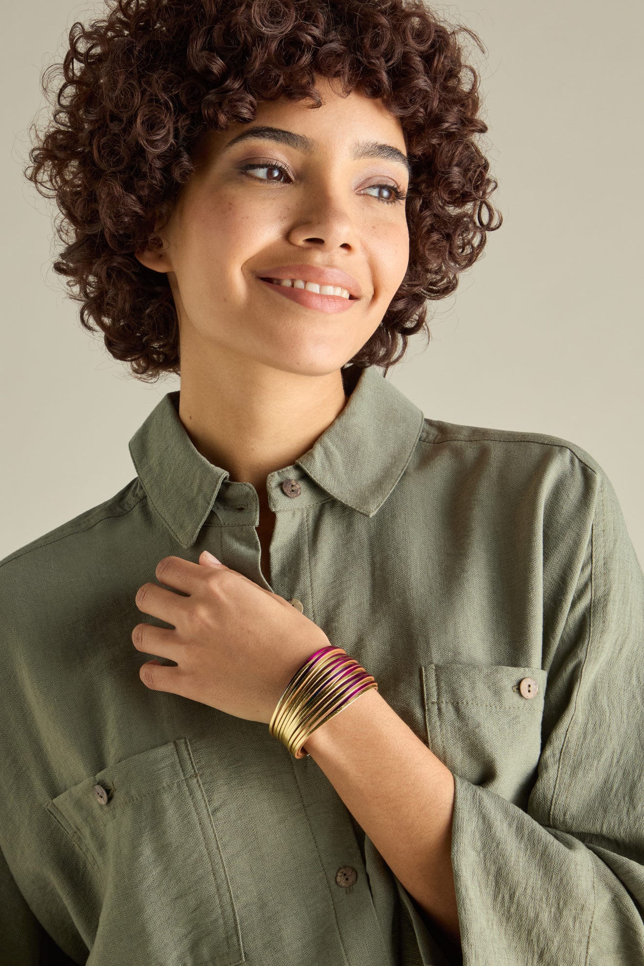 A person with curly hair wearing a green button-up shirt and several gold bracelets, including the Woven Metallic Bracelet featuring a twisted leatherette design and magnetic clasp, smiles while looking slightly to the side.