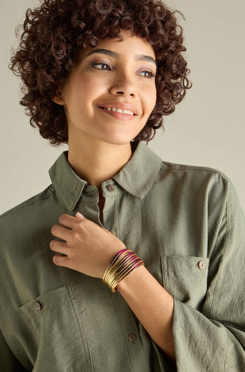 A person with curly hair wearing a green button-up shirt and several gold bracelets, including the Woven Metallic Bracelet featuring a twisted leatherette design and magnetic clasp, smiles while looking slightly to the side.