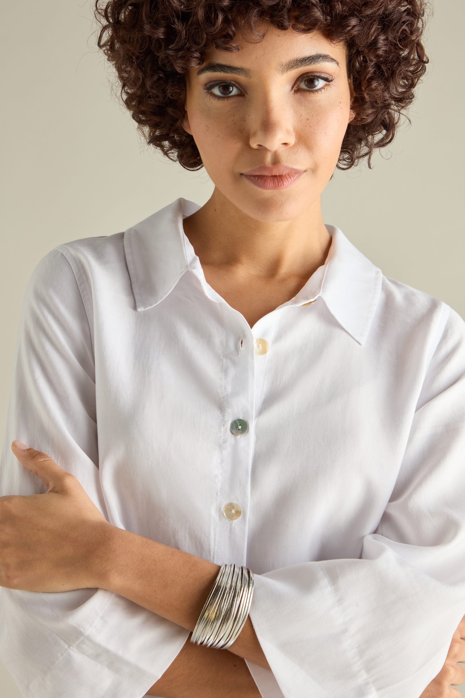 A person with curly hair and a white button-up shirt is wearing a Woven Metallic Bracelet featuring a magnetic clasp, posing for the camera with arms crossed.