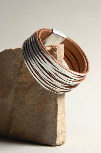 The Woven Metallic Bracelet, crafted with silver and brown leather and featuring a subtle metallic finish, is elegantly showcased on a rectangular stone slab.