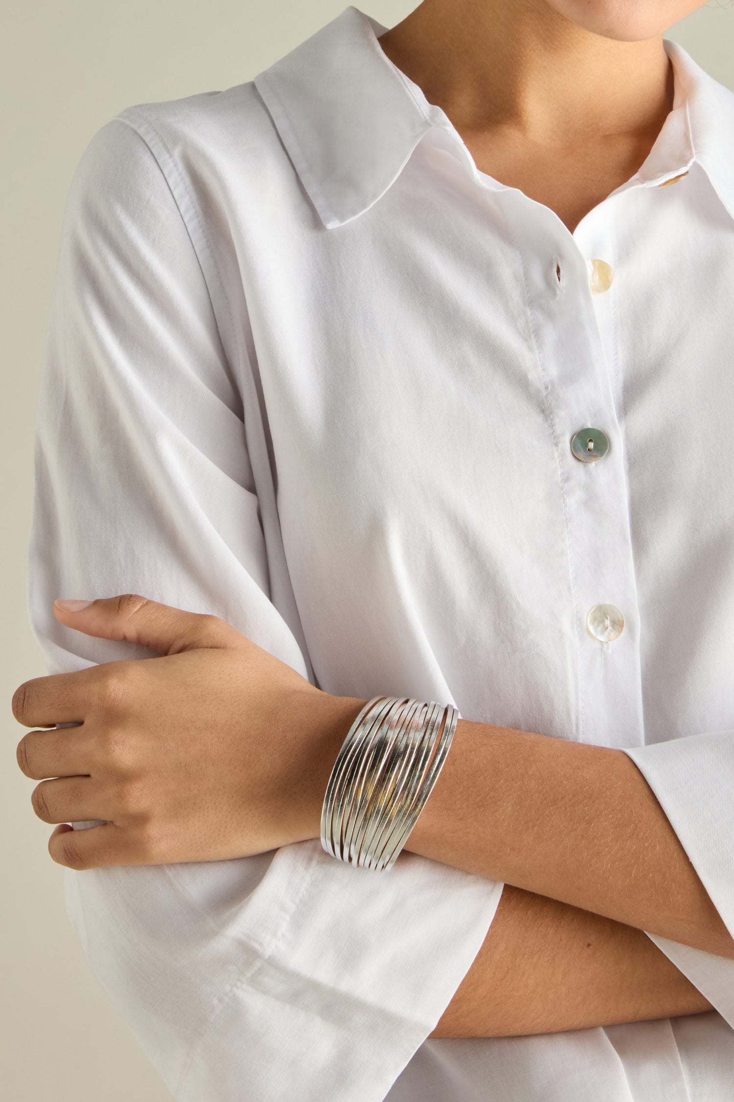 A person in a white blouse with arms crossed is wearing a stack of silver bangles interwoven with a Woven Metallic Bracelet.