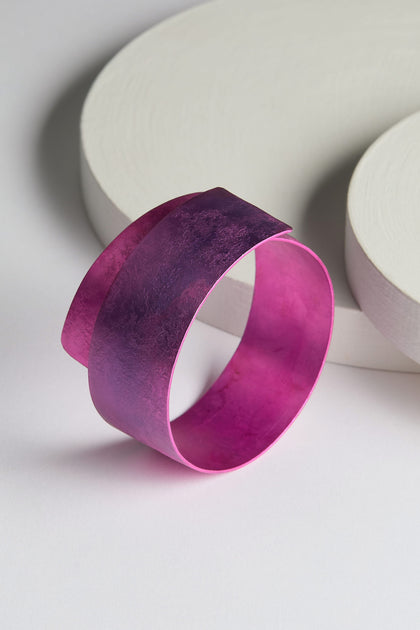 The Curved Bracelet, a wide, metallic piece in a striking purple hue with a textured surface, rests on a white background decorated with circular shapes, showcasing modern design in aluminum jewelry.