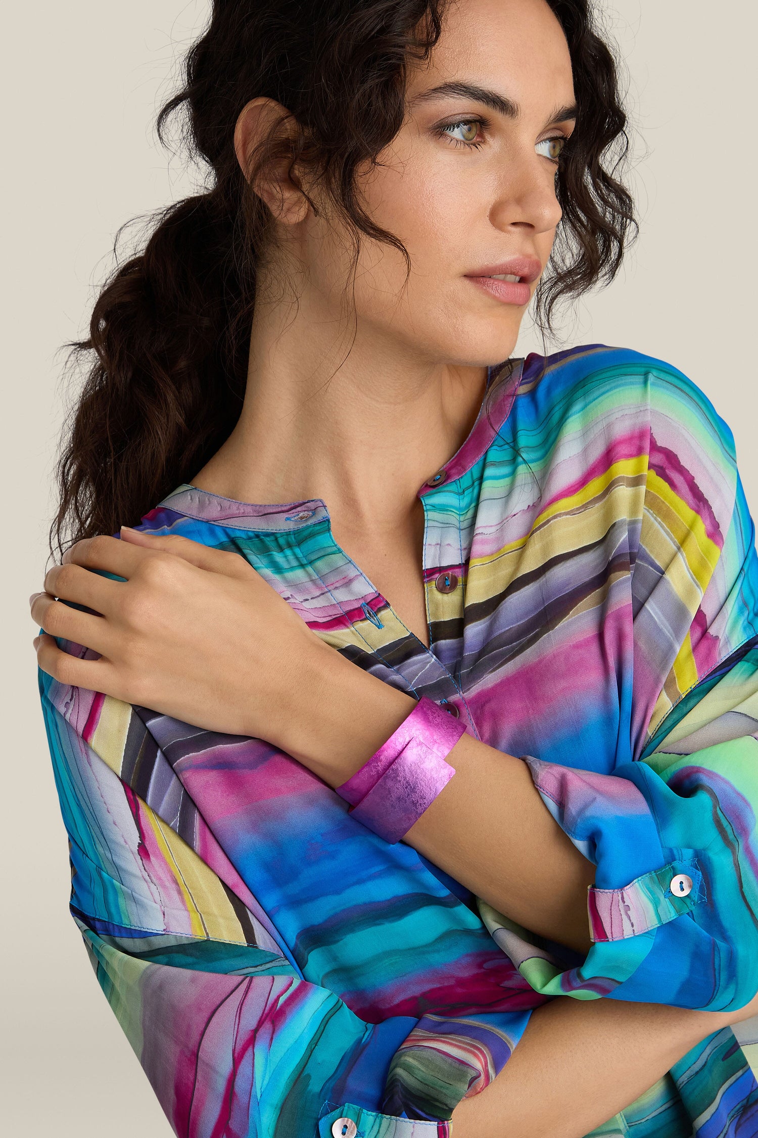 A person with wavy hair wears a colorful, striped top and a handcrafted Curved Bracelet, gazing to the side with one arm crossed over their chest.