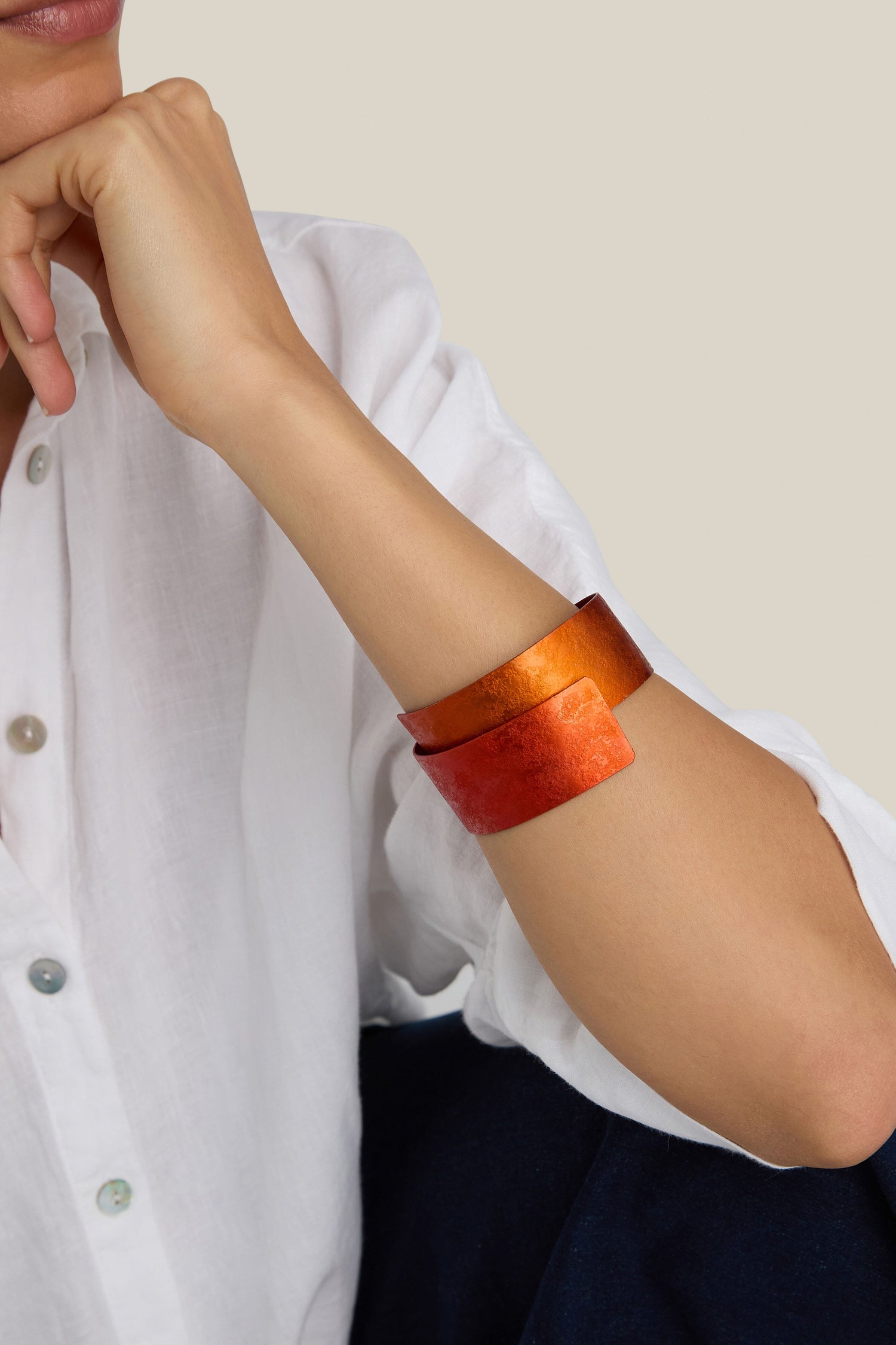 A person wearing a white shirt and a Curved Bracelet in an orange-red metallic finish, showcasing its modern design, rests their chin on their hand.