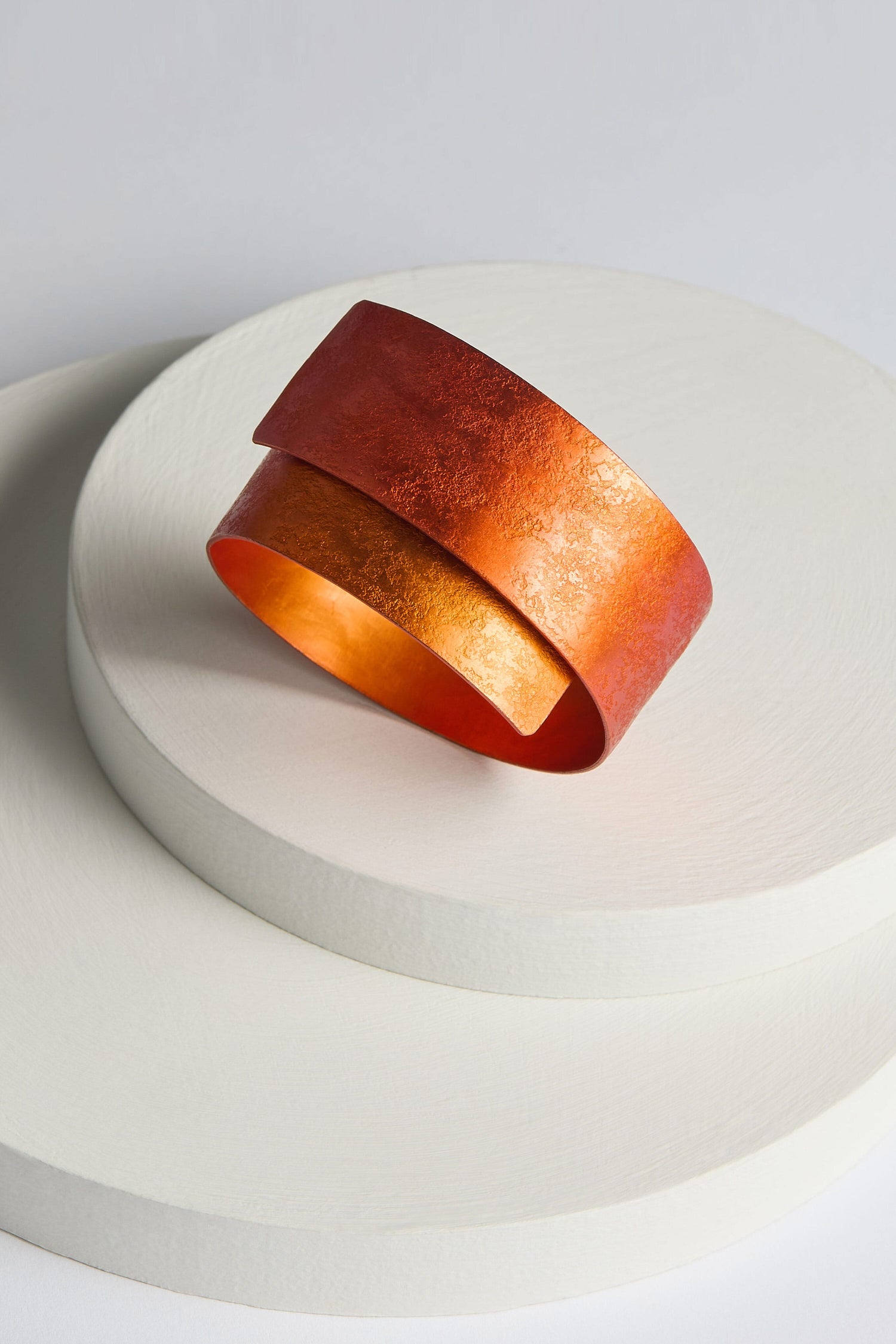 The Curved Bracelet, an orange-red aluminium jewellery cuff with a textured surface, is displayed on a round, white platform, showcasing its modern design against a minimalist background.