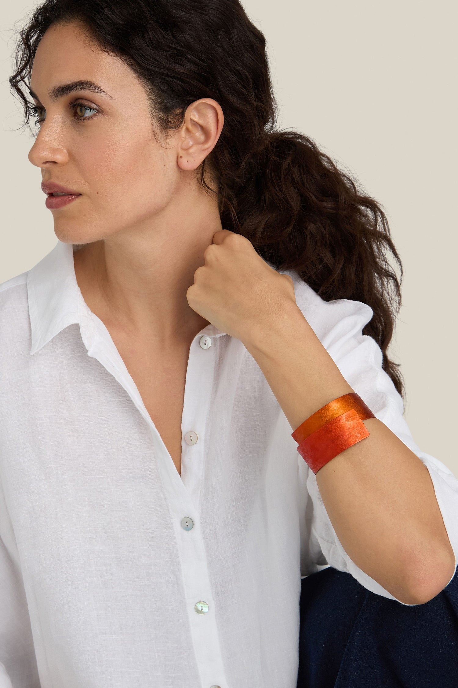 A person with dark hair pulled back, wearing a white button-up shirt and an orange-red Curved Bracelet, looks to the side against a light beige background.