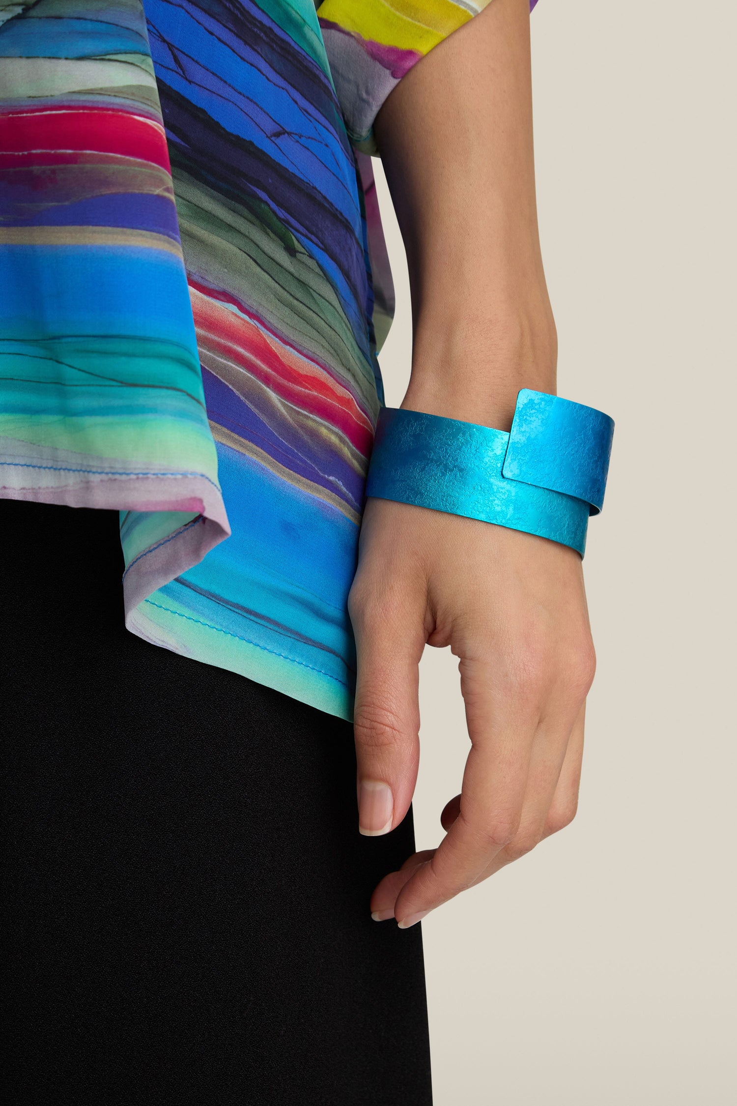 A person wearing a brightly colored top with stripes and an electric blue, handcrafted Curved Bracelet on their wrist. The person's hand is visible against a plain background, showcasing the modern design.