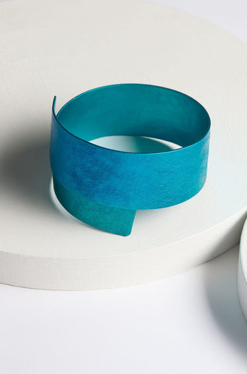 The Curved Bracelet, a wide, teal-colored aluminium accessory with a modern, minimalist design, is displayed on a white circular platform.