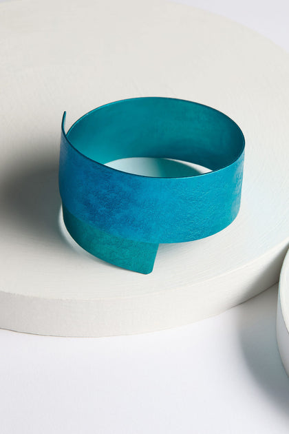 The Curved Bracelet, a wide, teal-colored aluminium accessory with a modern, minimalist design, is displayed on a white circular platform.