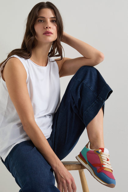 A woman sits confidently on a stool, wearing a white sleeveless top, dark blue pants, and the chic Andree Trainers made from recycled mesh. She gazes directly at the camera, effortlessly blending style with sustainability.