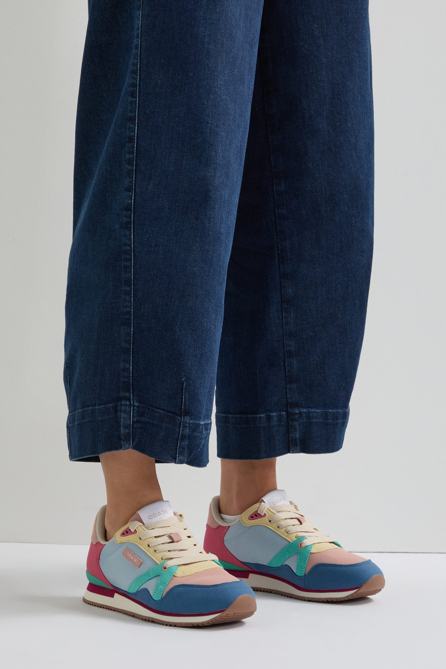 Standing on a bright surface, someone showcases blue jeans paired with Andree Trainers. These colorful sneakers feature pink, blue, and mint green accents and include recycled mesh details for an eco-conscious style statement.