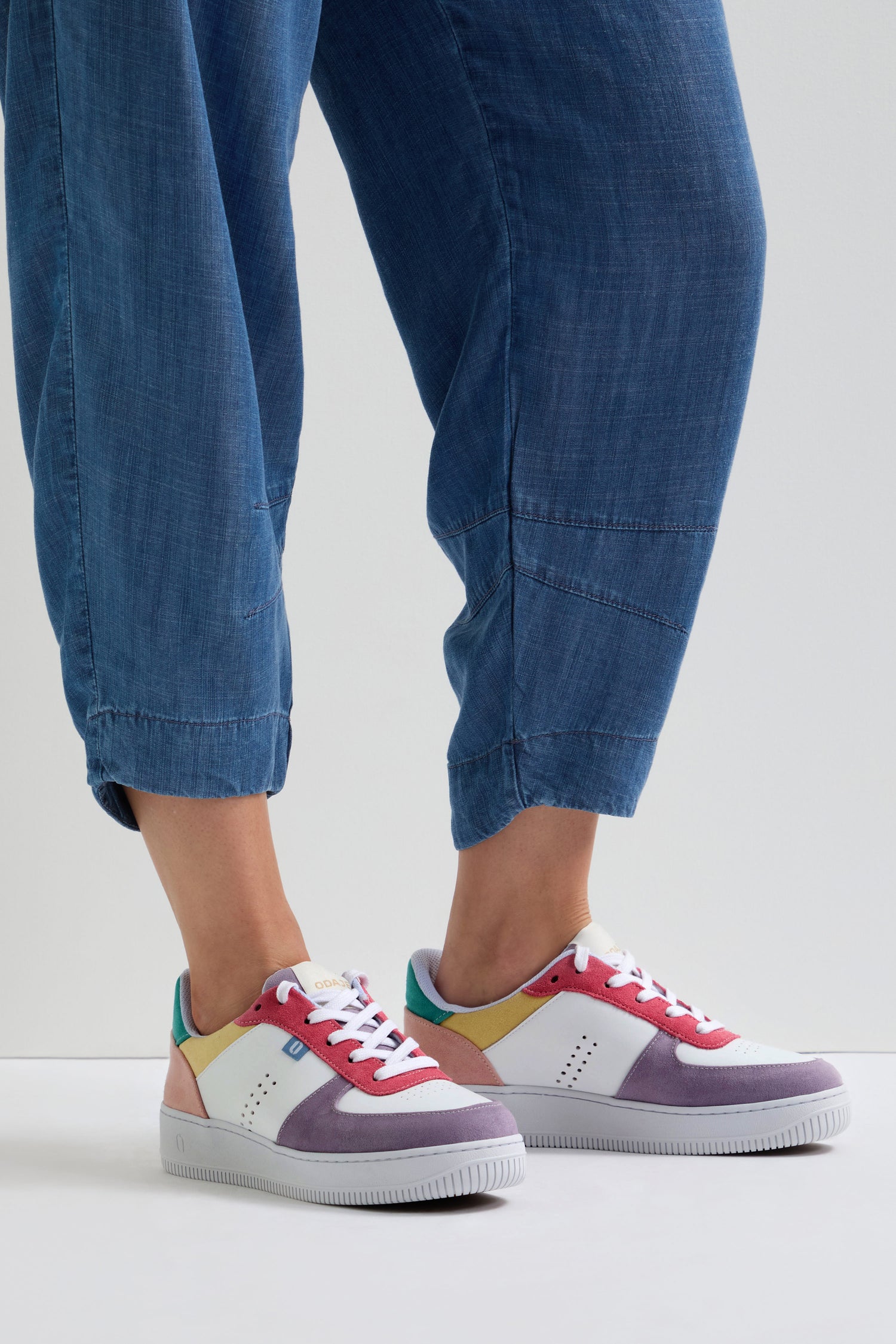 Against a white background, someone wearing blue pants confidently highlights the vibrant Maxence Trainers, made from vegan suede.