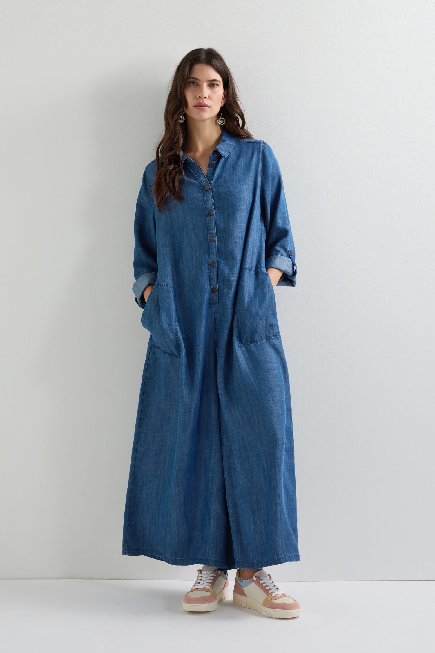 Person in a long blue denim dress and Nora Trainers stands against a white background.