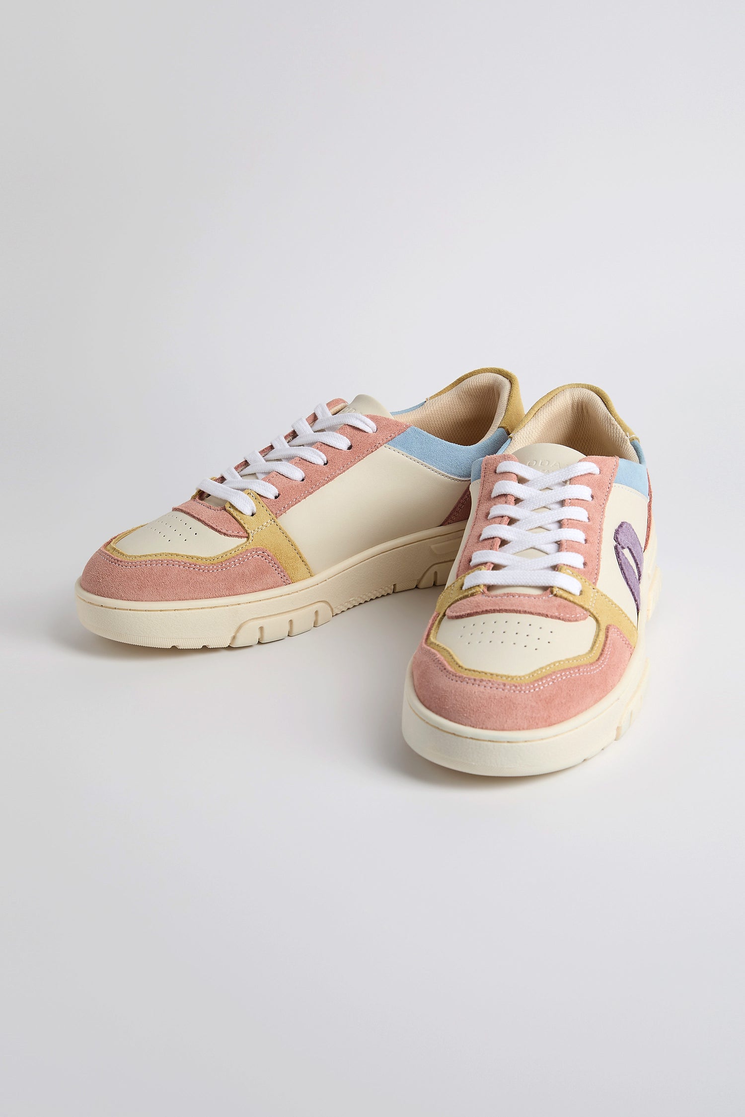 Nora Trainers, made from vegan suede, boast pastel pink, blue, and cream panels with white laces and beige soles. Set against a neutral backdrop, they reflect sustainable craftsmanship in every detail.