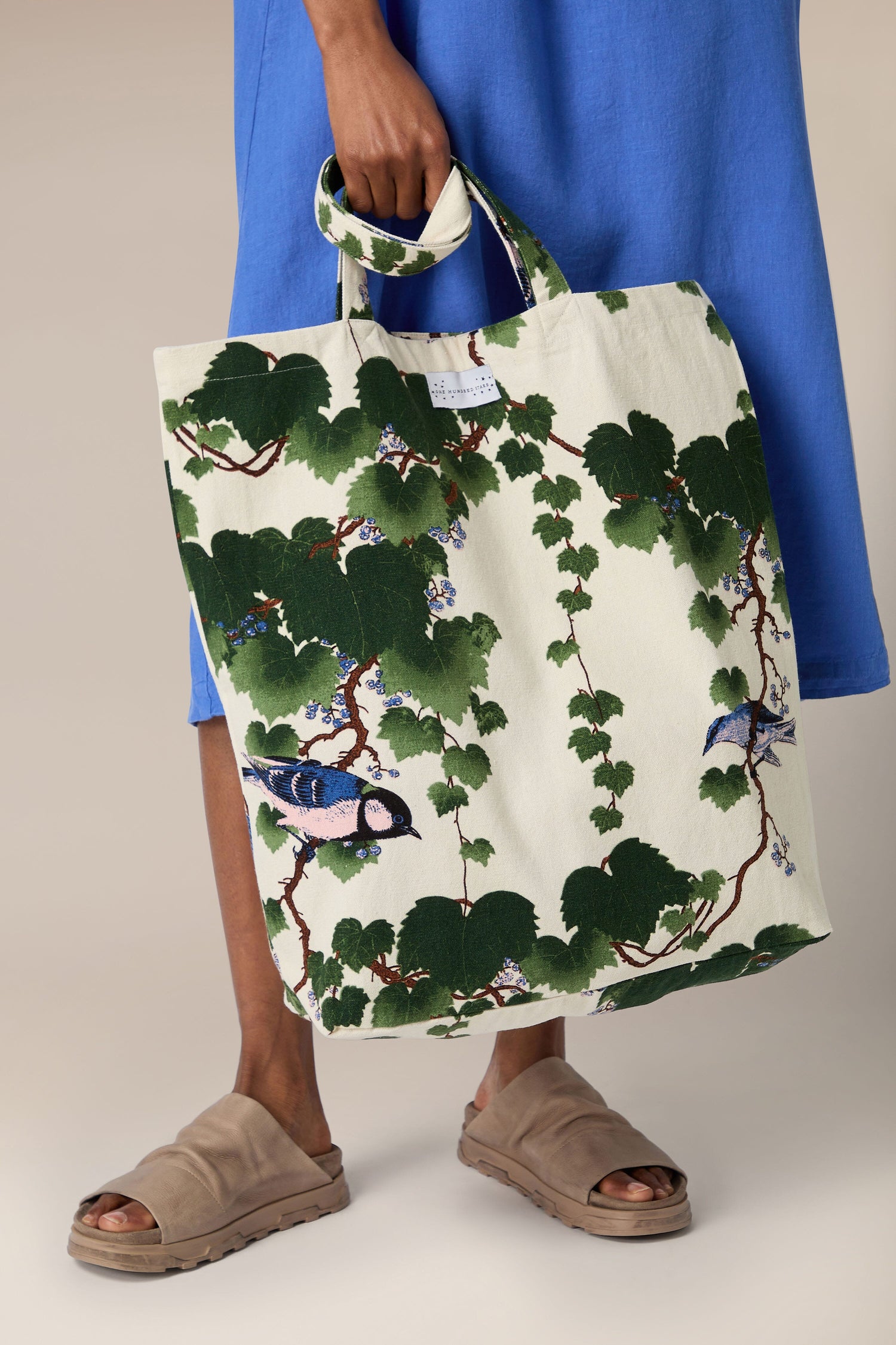 A person in a blue dress holding an Acer Canvas Bag with a leaf and bird pattern, standing in brown sandals.